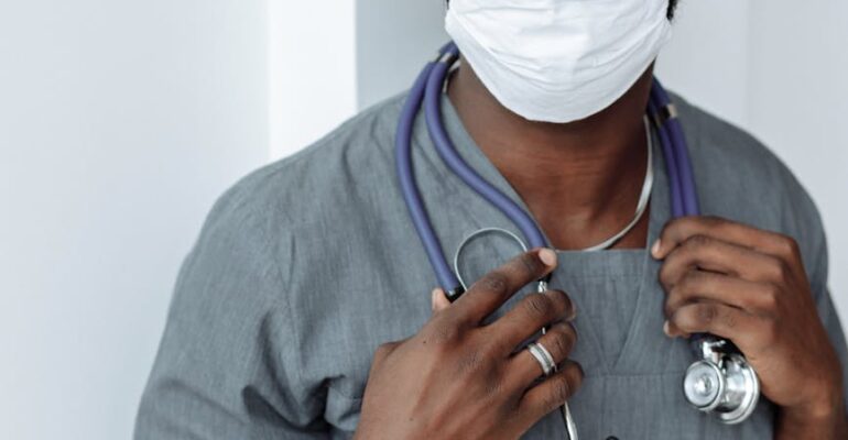 healthcare worker in gray scrubs suit with stethoscope on his neck wearing face mask while looking at the camera